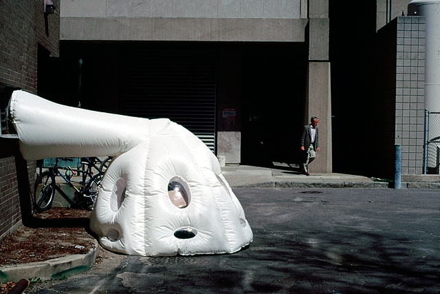 A photo of an artist made tent near a building entrance. The tent is made of white plastic, has round windows and a tube coming out of its top and going to the exhaust vent of the building. 
