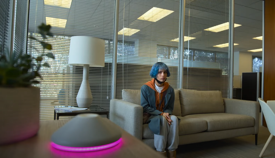 Still frame of Angela Childs--a light-skinned Black woman with blue hair--sitting in a waiting room. Angela is looking at Kimi, an Alexa-like smart speaker which sits in the foreground. Kimi is dark gray and shaped like a cone. In this photo, Kimi's base is lit up with pink lights, indicating that it is listening to its surroundings.