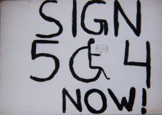 Handwritten protest sign from 1977, reads Sign 504 Now! The zero is shaped like a disability access symbol of a stick figure and wheelchair who is holding a sign.