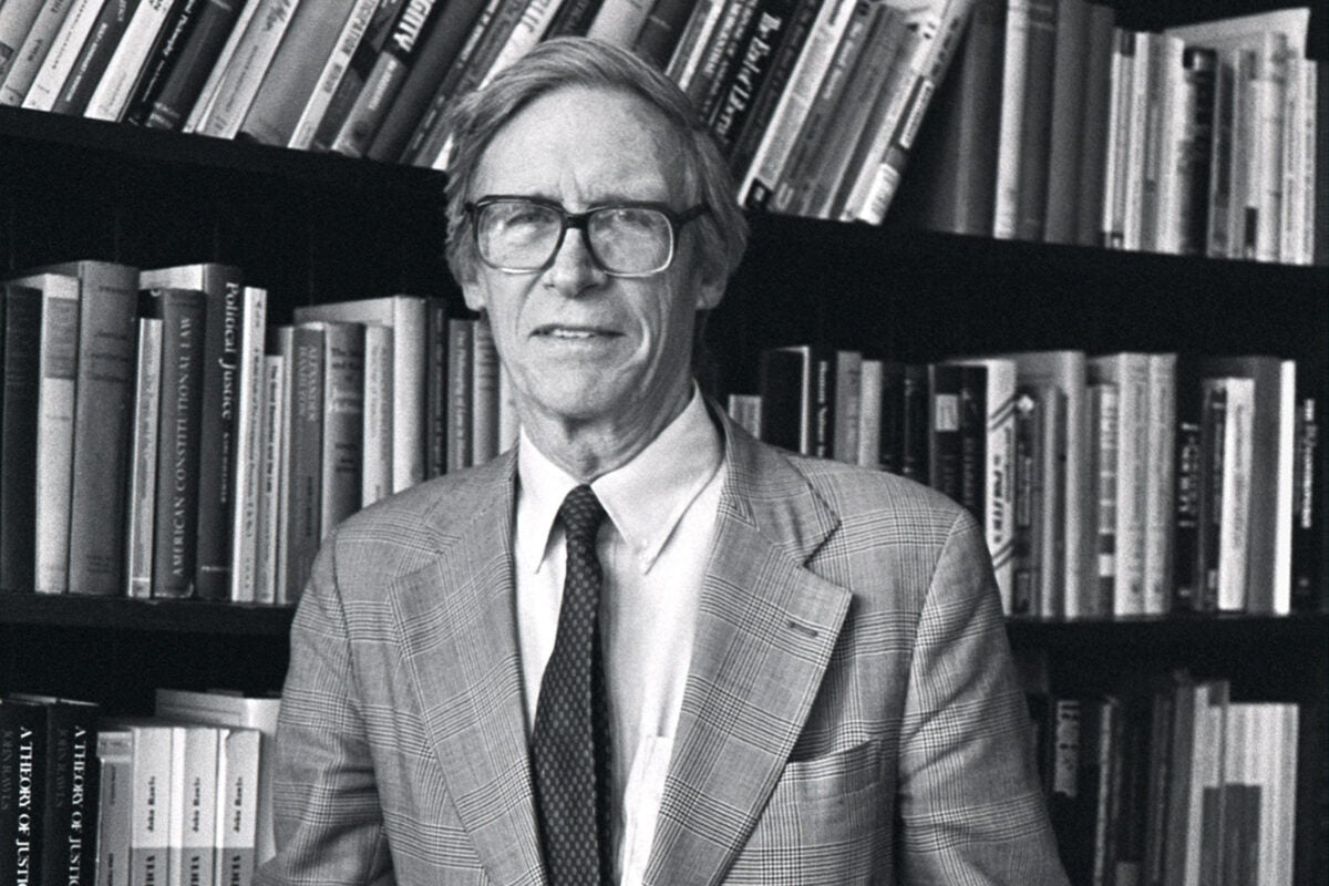 A black and white picture of John Rawls, an older white man with thick rimmed glasses, white hair, and a slight smile, in front of a shelf full of books with